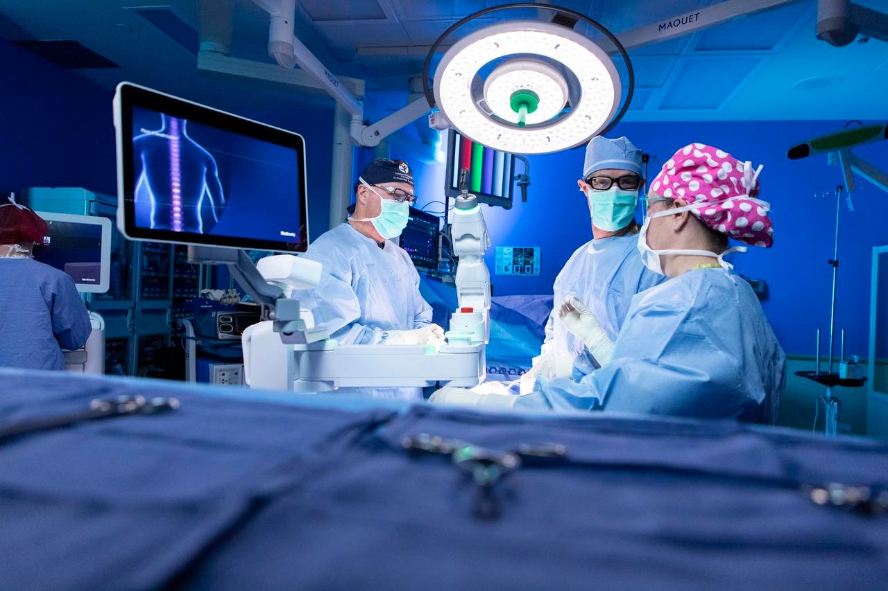 Three doctors in scrubs and masks stand in the operating room around a white surgical robot. There is a screen with a digital image of a patient’s spine.