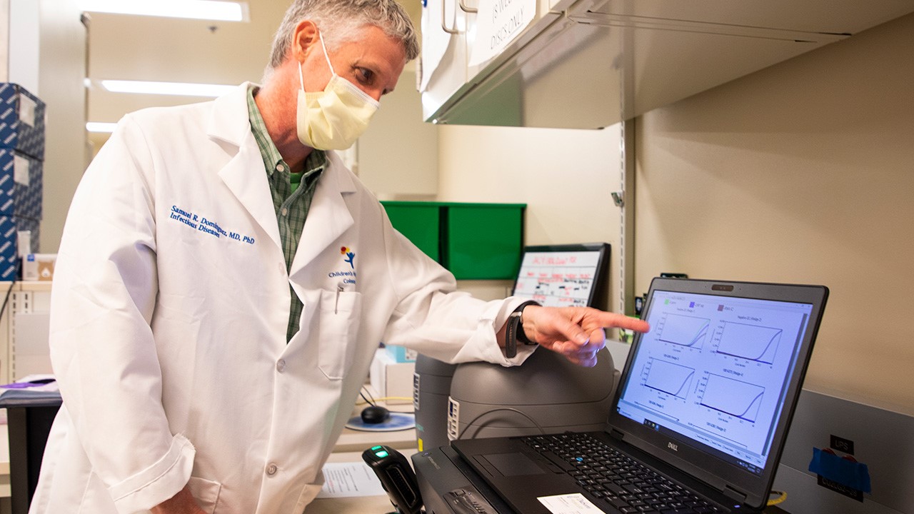 Samuel Dominguez, MD, PhD, wears a white lab coat and mask while pointing at data on a laptop in a laboratory setting.