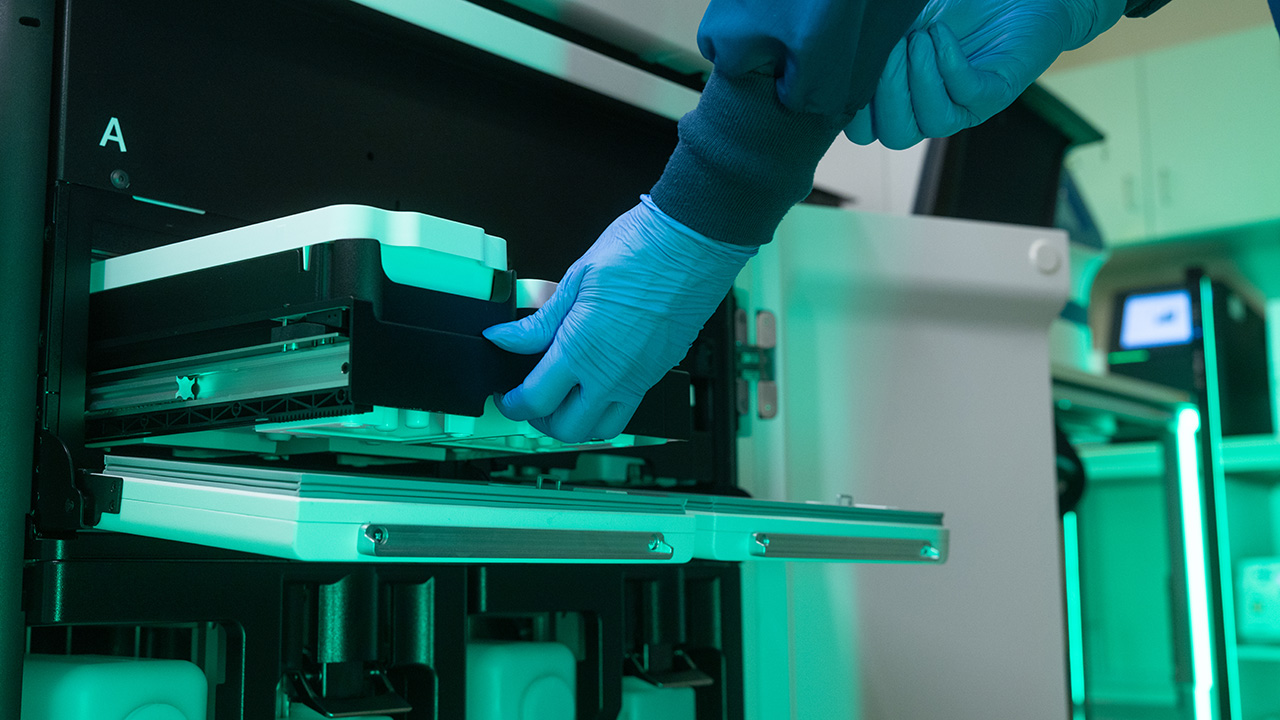  A person with gloved hands pushes a tray of genetic material into a machine for genetic sequencing. 