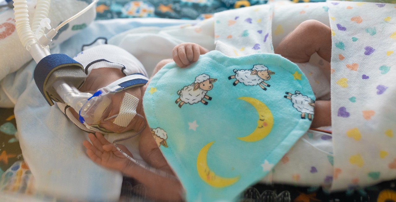 A baby in the neonatal intensive care unit lies in an isolette with a breathing tube while holding a felt heart.