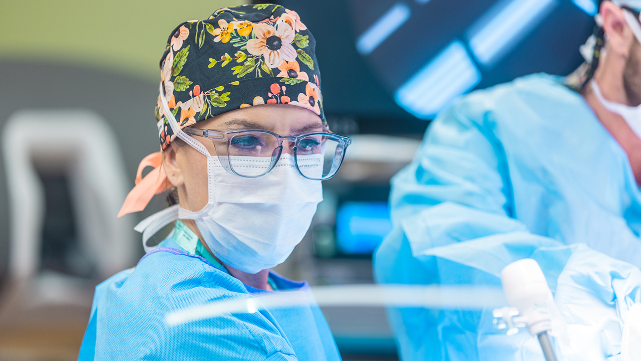 A doctor wears a flowered surgical cap, glasses, a face mask and scrubs while performing a surgery. 