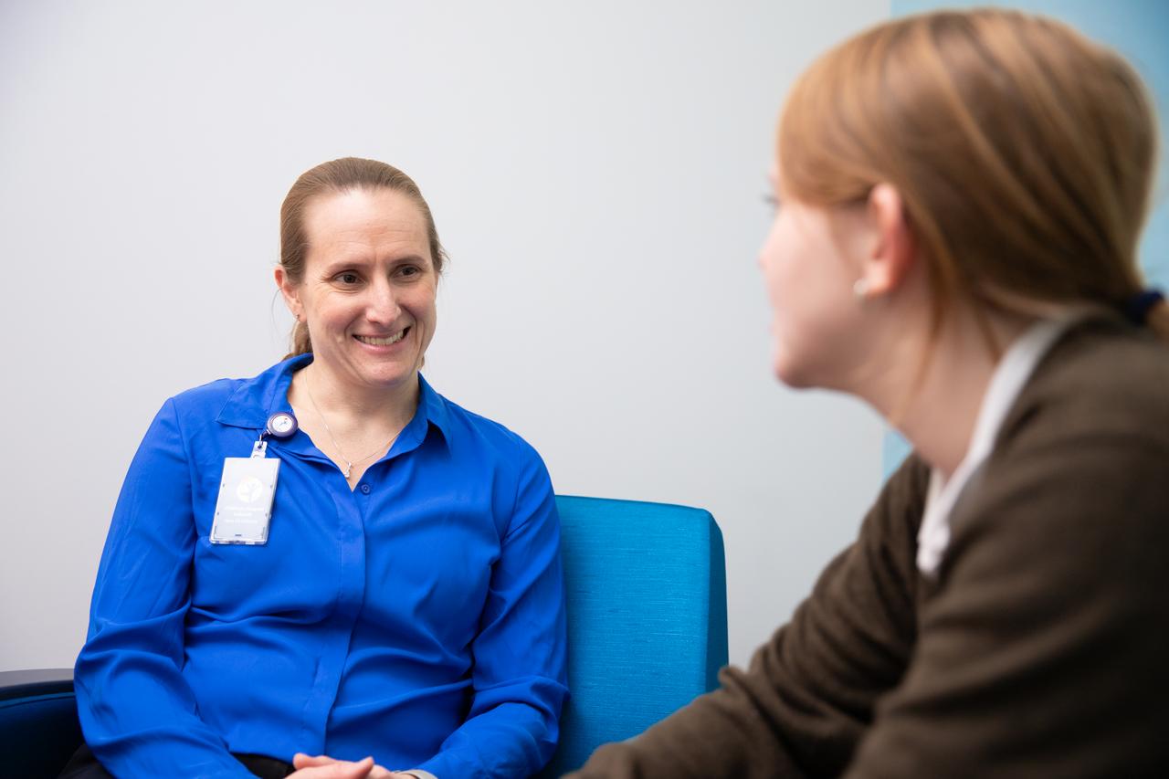 A teen sits in hospital room in a brown sweater and talks to a mental health care provider wearing a blue button-up shirt.