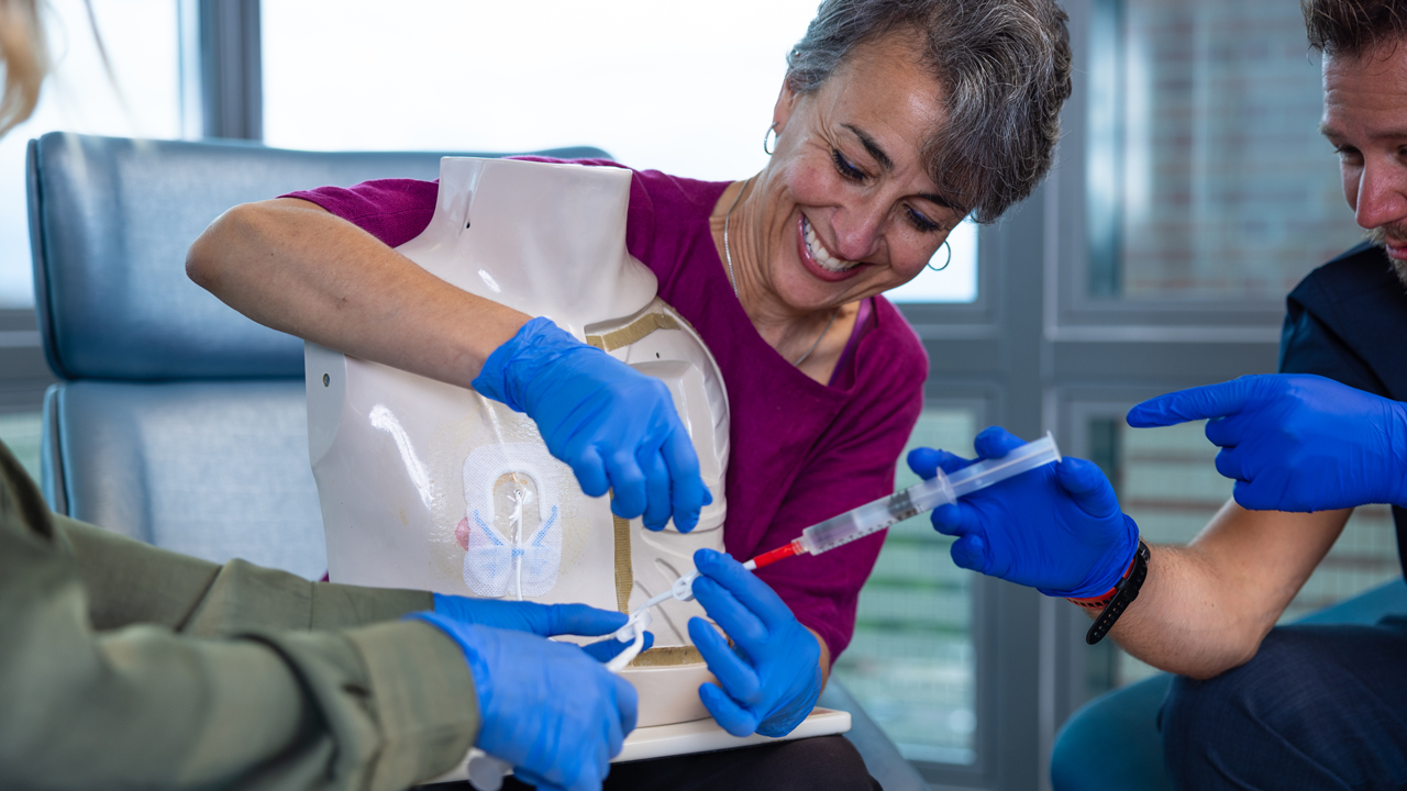 Three gloved providers practice using a syringe on a medical dummy.