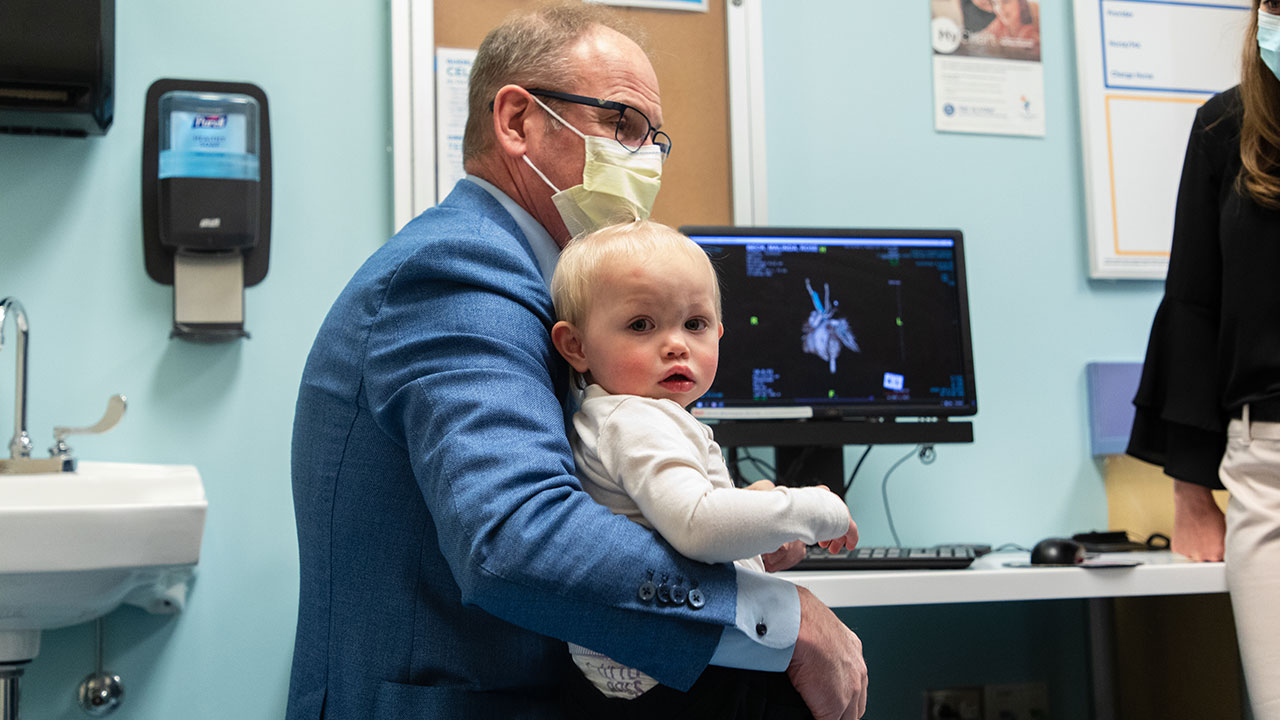 Pediatric cardiologist holds young patient on his lap with scans of a heart on the computer screen behind them.