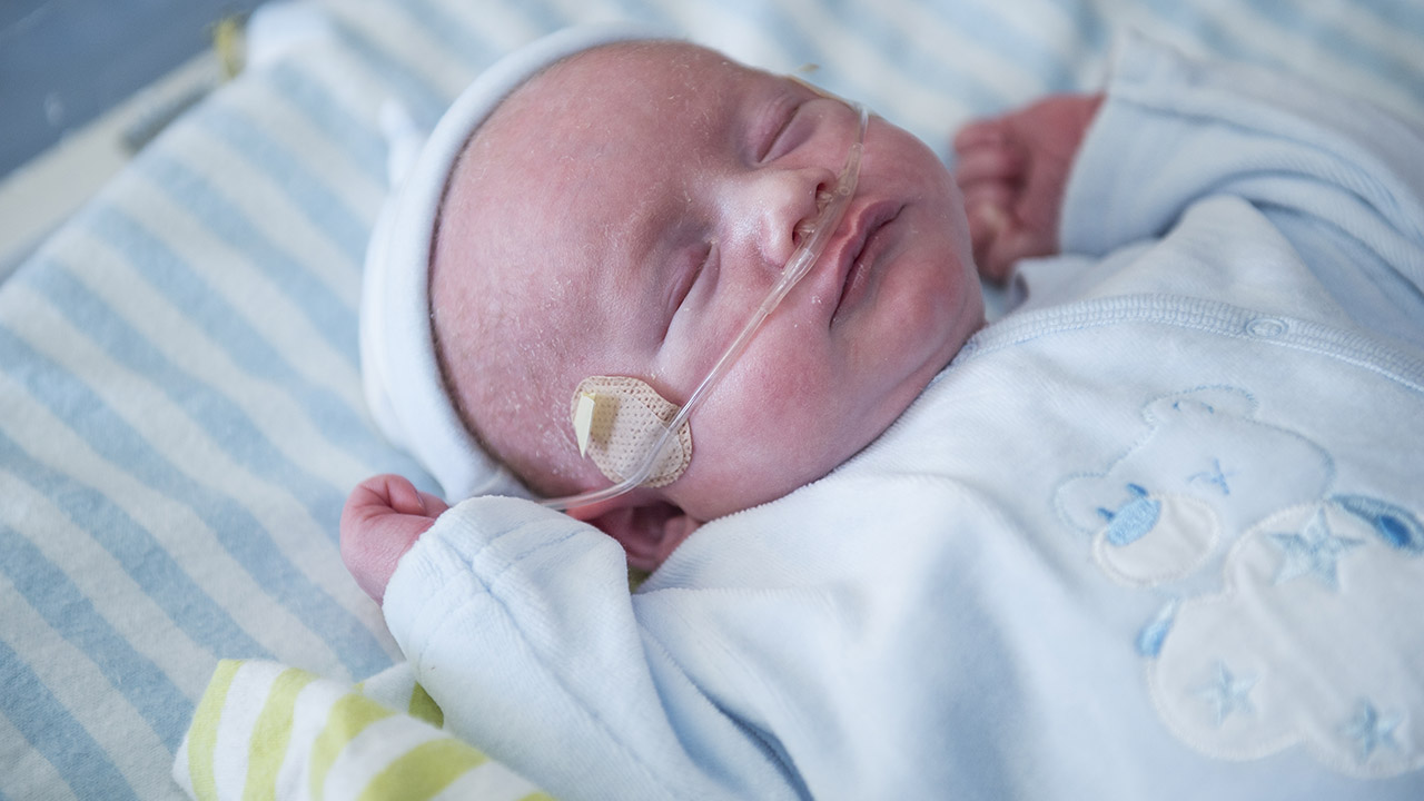 Close-up of sleeping baby with oxygen tube in nostrils. Dressed in blue, lying in the NICU.