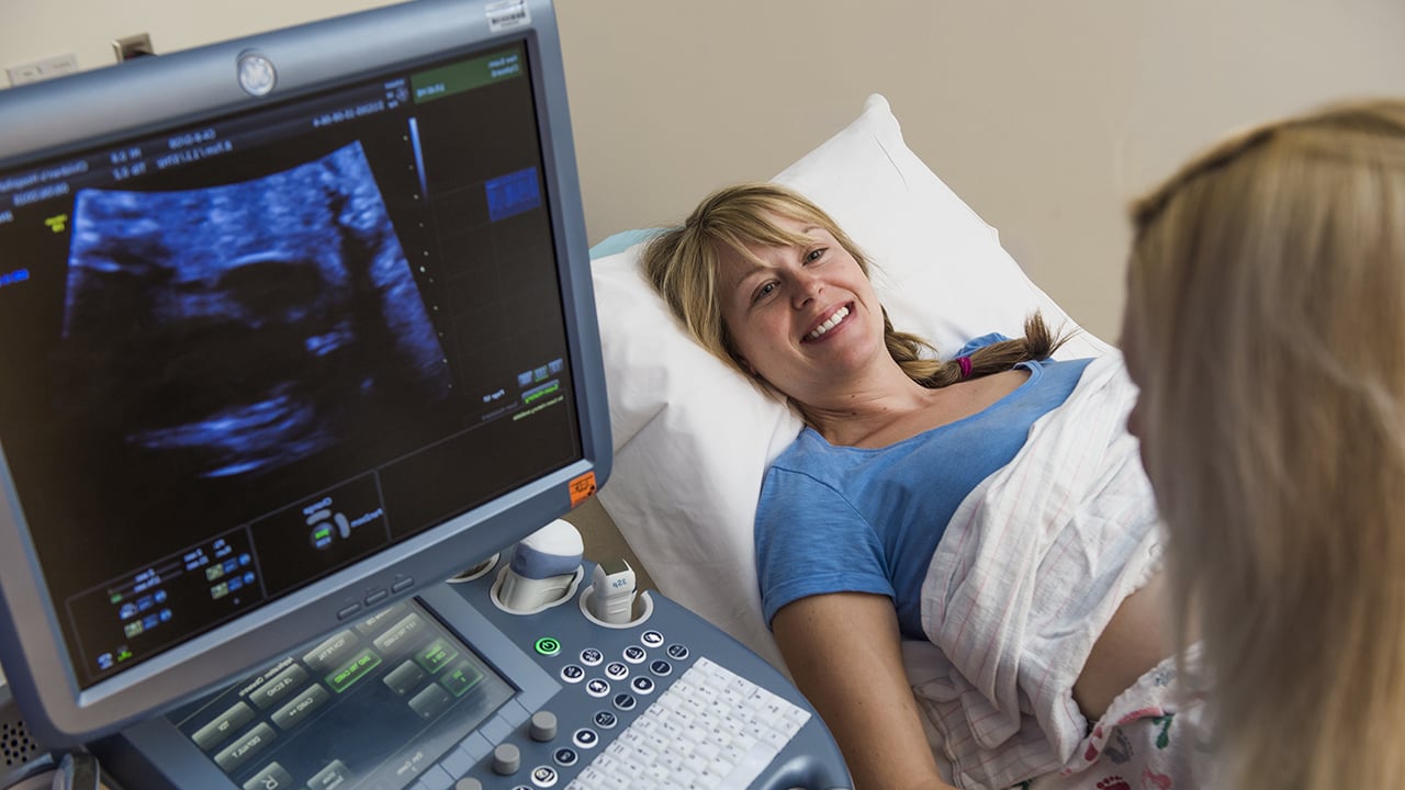 A smiling blond woman in a blue shirt lies on an exam table. Dr. Howley looks at a screen on a computer-like machine showing fetal ultrasound results. 