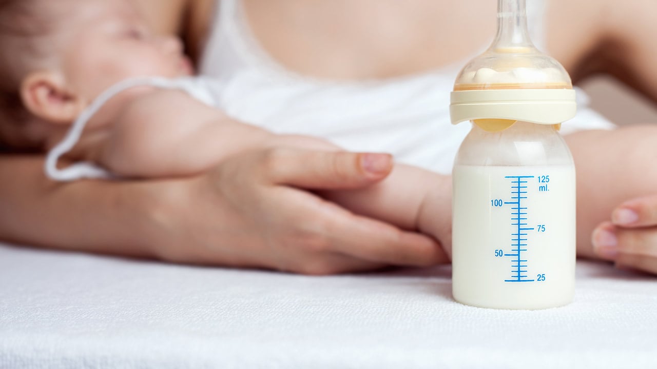 A close-up of a baby bottle full of breast milk on a table with the mom holding baby in the background.