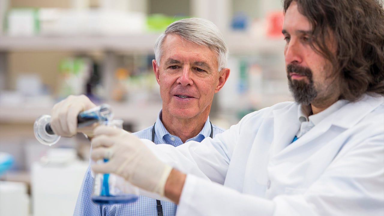 Dr. William Hay works on research in the lab, pouring blue liquid from one container to another.