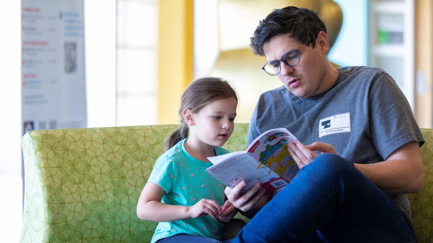 A parent wearing glasses and a white visitor’s badge flips through a book for kids while sitting with a child in the waiting room at a hospital or urgent care center.